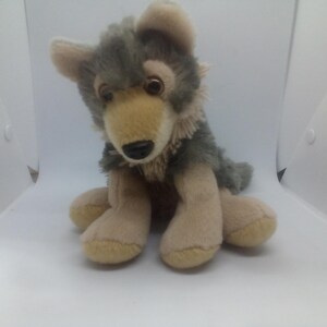 May include: A plush wolf toy featuring soft, shaggy fur in shades of grey and tan. The stuffed animal has large, dark brown eyes, pointed ears, and a black nose, depicted in a seated position with front paws resting on the ground.