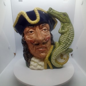 May include: A ceramic mug featuring a detailed face of a man wearing a tricorn hat with a yellow trim. A green alligator is perched on the side. The mug has a white ruffled collar and a yellow and green jacket.