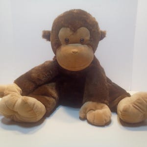 May include: A plush monkey toy featuring dark brown fur on the body and head, with lighter tan fabric on the face, hands, and feet. The stuffed animal has round ears, dark eyes, and a soft, seated posture with outstretched limbs and padded paws.