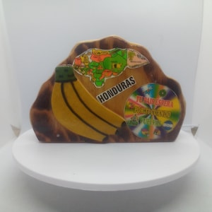 May include: A wooden napkin holder featuring a painted yellow banana, a map of Honduras, and a circular graphic with the text "TU PAIS ESPERA POR TI CUANDO VUELVAS". The wood has a natural brown finish with a contoured, irregular top edge design.