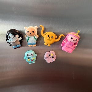 May include: A collection of colorful Adventure Time character magnets. The magnets include Finn, Jake, Princess Bubblegum, Marceline, BMO, and others. Each magnet is a unique, handcrafted design, perfect for decorating a refrigerator or locker.
