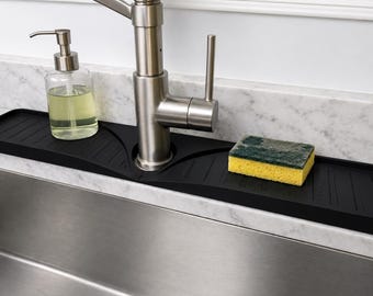 Könnte beinhalten: Eine schwarze Silikon-Spritzschutzmatte für Spülbeckenarmaturen mit rechteckiger Form und erhöhten Ablaufrillen. Die Matte verfügt über einen mittigen Ausschnitt für den Wasserhahn, einen transparenten Seifenspender und einen gelb-grünen Schwamm.