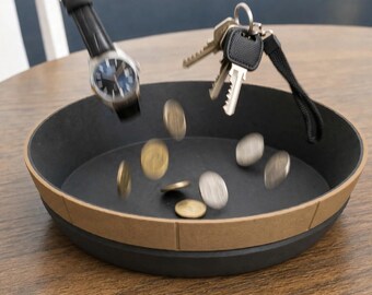 Könnte beinhalten: Eine runde, schwarze Ablageschale mit einem beigefarbenen Außenrand. Eine Armbanduhr, ein Schlüsselbund aus Metall mit schwarzem Band sowie mehrere goldene und silberne Münzen sind in der Luft zu sehen, wie sie in die Schale fallen.