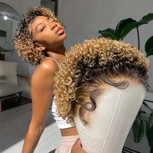May include: A curly wig featuring a dark brown root transition into bright blonde highlights. The hair is styled in tight, defined ringlets with a lace front hairline that mimics a natural growth pattern, displayed on a white mannequin head.
