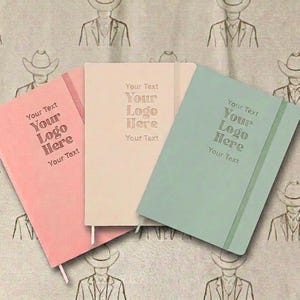 Peut inclure: Trois carnets à couverture rigide en rose, beige et vert clair, chacun doté d'une bande élastique de fermeture et du texte en relief "Your Text Your Logo Here Your Text" sur la couverture avant. Les carnets sont disposés côte à côte.