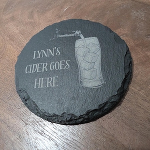 May include: A round, dark grey slate coaster with a rough, natural edge. The surface features an engraved illustration of a glass filled with ice cubes being poured from a bottle, alongside the text, LYNN'S CIDER GOES HERE, in a clean font.