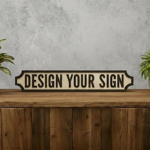 Op de afbeelding: Een horizontaal rechthoekig bord met een donkerbruine rand en een crèmekleurige achtergrond. De tekst "DESIGN YOUR SIGN" staat in dikke, donkerbruine, schreefloze hoofdletters in het midden van het bord, dat is voorzien van decoratieve uiteinden.