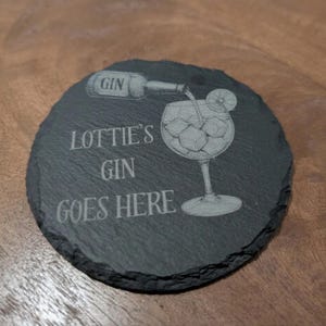 May include: A round, dark gray slate coaster with a rough, uneven edge. It features an engraved illustration of a gin bottle pouring into a glass filled with ice and a lime slice, with the text LOTTIE'S GIN GOES HERE etched in the center.