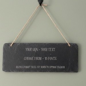May include: A rectangular black slate sign with white text, suspended by a natural twine cord. The sign reads "YOUR SIGN - YOUR TEXT", "CHOOSE FROM - 10 FONTS", and "RECYCLED ROOF TILES, CUT DOWN TO APPROX 25x10cm". The sign hangs against a muted green wall.