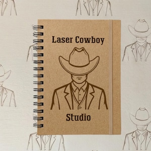 Peut inclure: Un carnet à spirales avec une couverture beige présentant un motif marron foncé d'une personne portant un chapeau de cowboy et un costume. Le texte Laser Cowboy est imprimé au-dessus et le mot Studio est imprimé en dessous.