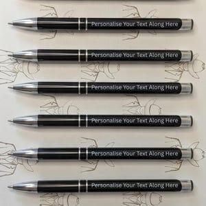 May include: Six black ballpoint pens with silver-toned metal tips, clips, and accents. Each pen features the white printed text "Personalise Your Text Along Here" on the barrel. The pens are arranged horizontally in a parallel stack against a light background.