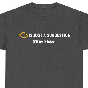 May include: Dark grey t-shirt featuring the text "IS JUST A SUGGESTION (I'll fix it later)" and a yellow engine light icon. The shirt is made of a soft material and has a crew neck.