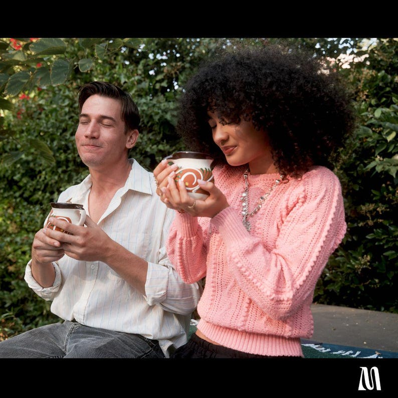 May include: Two people are shown enjoying a warm drink from ceramic mugs. The mugs have a brown and white design. The person on the right is wearing a pink sweater and the person on the left is wearing a white button-down shirt.