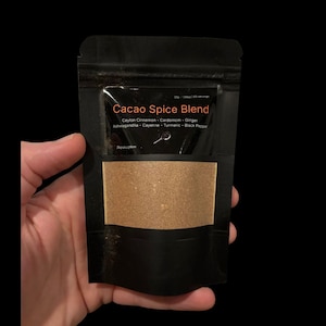 May include: A black pouch of Cacao Spice Blend. The pouch, held in a hand, has a clear window revealing the brown powder. Text on the pouch reads "Cacao Spice Blend" and lists ingredients including Ceylon Cinnamon and Cardamom.