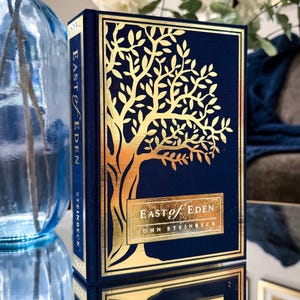 May include: A hardcover book titled East of Eden by John Steinbeck with a deep navy blue cover. The front features a large, intricate gold foil tree design and a rectangular gold foil label with the title and author name printed in white.