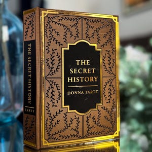 May include: A hardcover book titled "The Secret History" by Donna Tartt. The book has a brown cover with gold accents and a decorative leaf pattern. The title is in a black rectangle with gold trim.