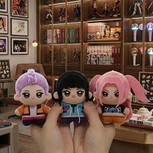 Pu&ograve; includere: Tre bambole di peluche con caratteristiche in stile anime. Le bambole hanno acconciature e abiti diversi, tra cui uno chignon viola, capelli neri e codini rosa. Le bambole sono tenute davanti a uno sfondo con scaffali di libri e statuette.