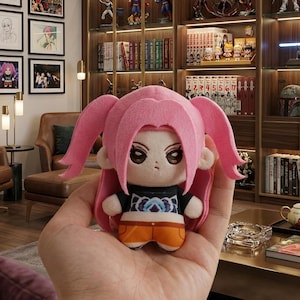 Pu&ograve; includere: Una bambola di peluche con capelli rosa e un top nero con un disegno blu e bianco. La bambola ha pantaloncini arancioni ed &egrave; tenuta in mano. La bambola &egrave; un personaggio di una serie anime.
