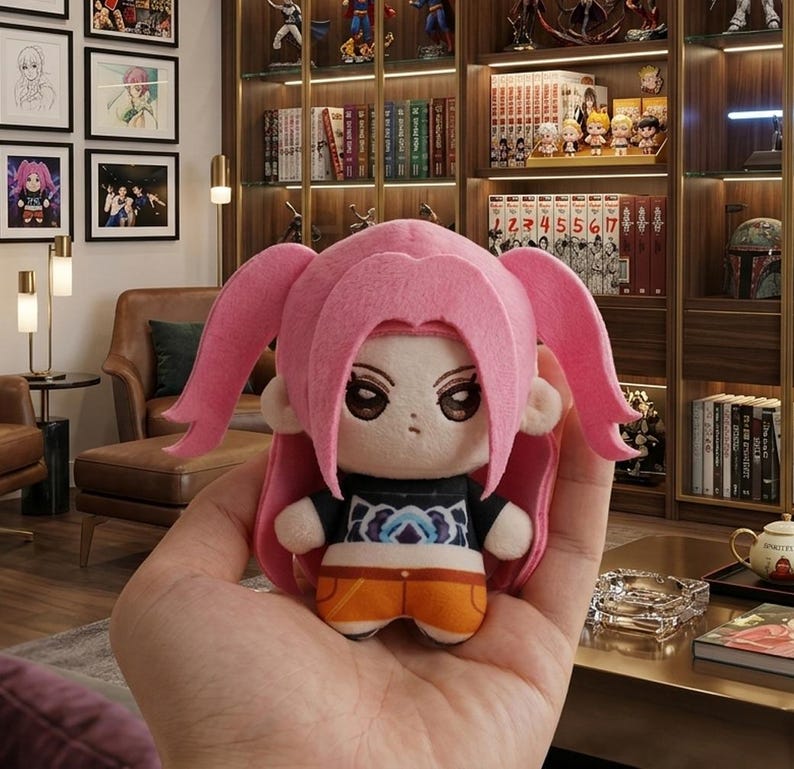 Pu&ograve; includere: Una bambola di peluche con capelli rosa e un top nero con un disegno blu e bianco. La bambola ha pantaloncini arancioni ed &egrave; tenuta in mano. La bambola &egrave; un personaggio di una serie anime.