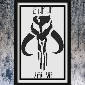 May include: A framed cross-stitch pattern featuring a black Mythosaur skull emblem centered on a white background. The design includes stylized text above and below the central icon, all enclosed within a simple black rectangular frame.