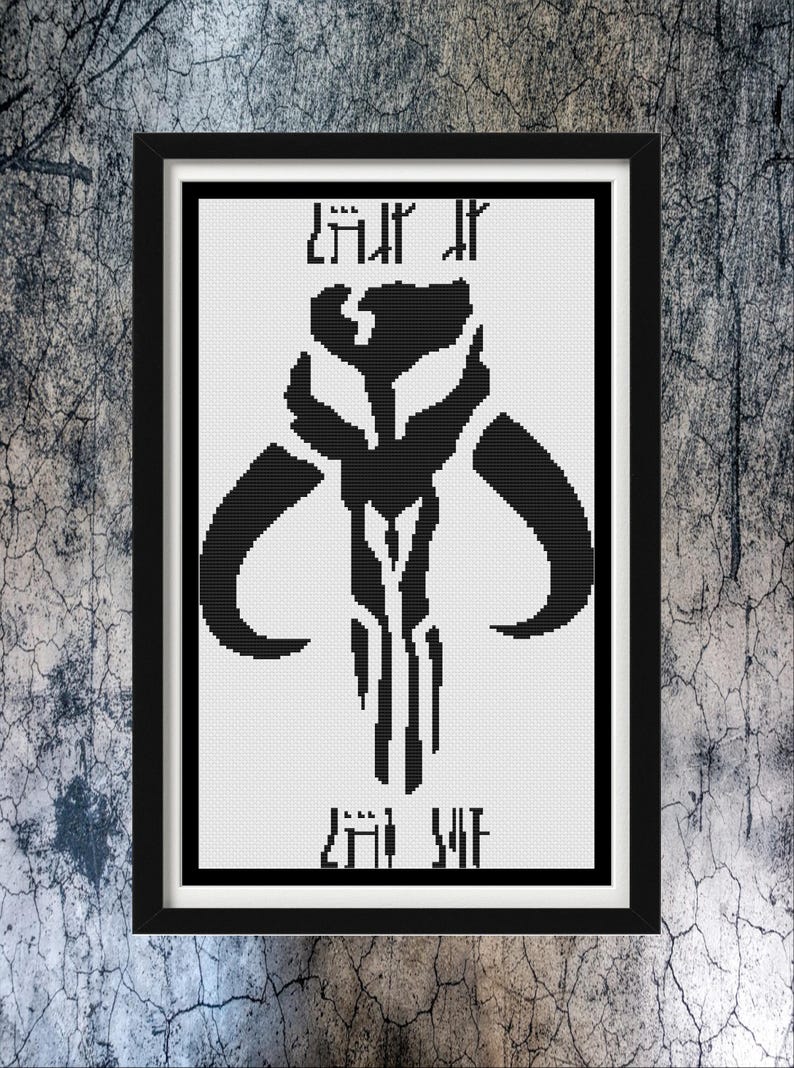 May include: A framed cross-stitch pattern featuring a black Mythosaur skull emblem centered on a white background. The design includes stylized text above and below the central icon, all enclosed within a simple black rectangular frame.