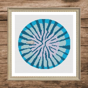 May include: A framed cross-stitch artwork with a circular design. The design features a central light purple area with radiating lines in shades of blue and teal. The artwork is in a light brown wooden frame, set against a wood-grain background.