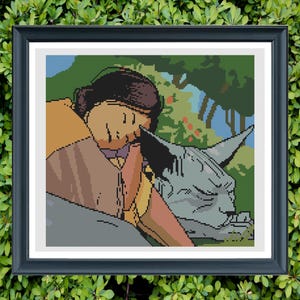 May include: Cross-stitch art in a black frame. The artwork depicts a person resting their head on a gray, stylized cat statue. The background features green foliage and blue sky, creating a serene, pixelated scene.