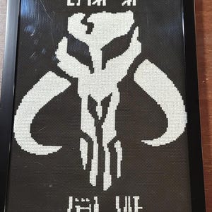 May include: A framed cross-stitch artwork featuring a white Mythosaur skull emblem centered on a black fabric background. The design includes stylized Aurebesh text positioned both above and below the central skull icon, all encased in a black frame.