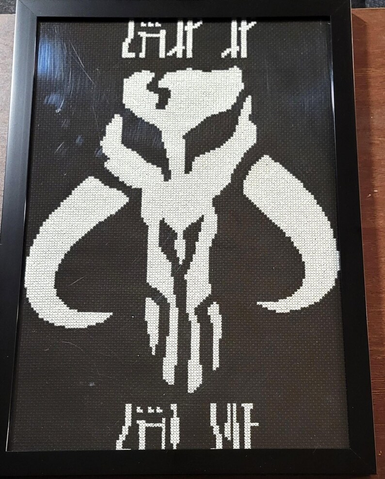 May include: A framed cross-stitch artwork featuring a white Mythosaur skull emblem centered on a black fabric background. The design includes stylized Aurebesh text positioned both above and below the central skull icon, all encased in a black frame.