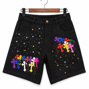 May include: Black denim shorts with colorful cross embroidery and scattered jewel accents. The shorts are hanging on a wooden hanger. The crosses are in various colors, including purple, yellow, and pink.