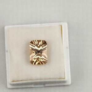 May include: A rectangular, faceted gemstone with a warm, peach-toned color. The gem is set in a white display case, showcasing its intricate internal structure and brilliance. The gem's cut creates a play of light.