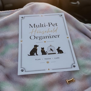 May include: A white rectangular Multi-Pet Household Organizer planner with black text and gold accents. The cover features silhouettes of a dog, cat, bird in a cage, and rabbit, with the text PLAN • TRACK • CARE and a small gold bone-shaped pin.