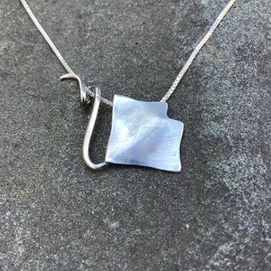 May include: A silver necklace with a unique, abstract pendant. The pendant is a flat, irregular shape with a brushed finish. The chain is a delicate, box chain.