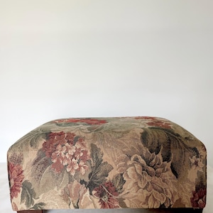 May include: A rectangular ottoman upholstered in a woven tapestry fabric featuring a dense floral pattern. The design displays large blossoms in shades of deep red, muted pink, and earthy green against a tan background, supported by dark brown feet.