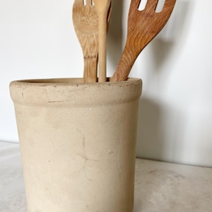 May include: A beige ceramic utensil holder containing wooden cooking utensils. The holder is cylindrical with a slightly textured surface. The wooden utensils include a fork, a spoon, and a slotted spoon. The utensils are various shades of brown.