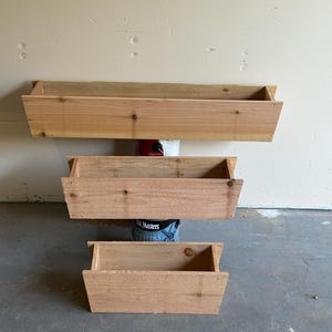 Könnte beinhalten: Drei rechteckige Holzpflanzkästen unterschiedlicher Größe sind vertikal gestapelt. Die Pflanzkästen sind aus hellfarbenem Holz gefertigt und haben ein einfaches, offenes Design. Sie sind an einer hellfarbigen Wand montiert, was auf ein Garten- oder Außendekorationsthema hindeutet.
