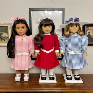 May include: Three dolls dressed in vintage-style dresses. The dolls have long hair and are wearing dresses in pink gingham, red, and blue gingham. Each doll has a white belt and black shoes. One doll wears a hat with flowers.
