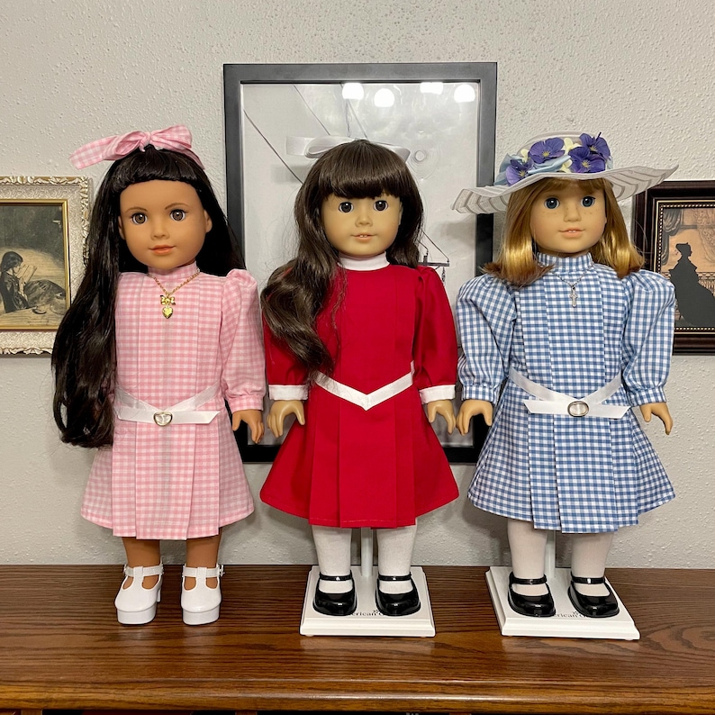 May include: Three dolls dressed in vintage-style dresses. The dolls have long hair and are wearing dresses in pink gingham, red, and blue gingham. Each doll has a white belt and black shoes. One doll wears a hat with flowers.