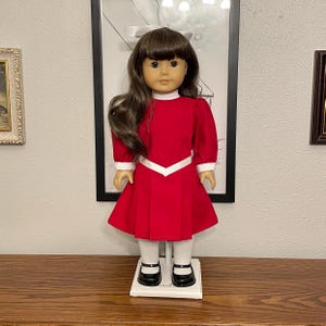 May include: A doll with brown hair and brown eyes wearing a red dress with white trim, white socks, and black shoes. The doll is standing on a white stand. The doll is in front of a framed picture.