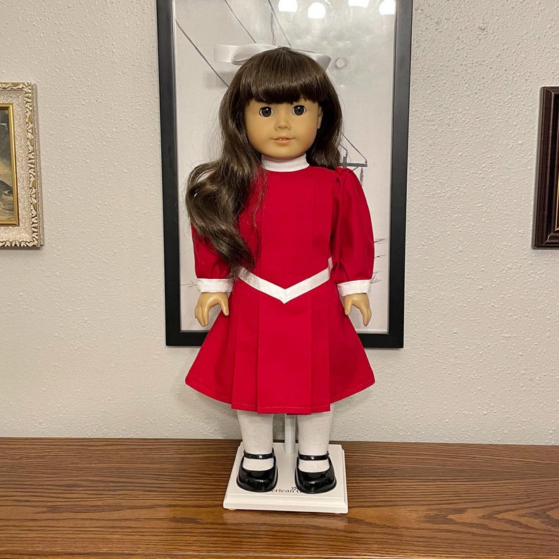 May include: A doll with brown hair and brown eyes wearing a red dress with white trim, white socks, and black shoes. The doll is standing on a white stand. The doll is in front of a framed picture.
