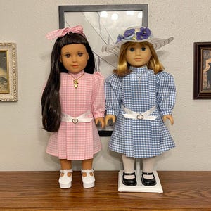May include: Two dolls dressed in vintage-style gingham dresses. One doll wears a pink dress with a matching bow, the other a blue dress with a floral hat. Both dolls have white belts and shoes. The dolls are displayed on a wooden surface.