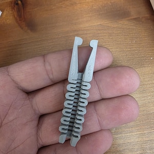 Puede incluir: Un clip de plástico gris impreso en 3D, sostenido en una mano. El clip tiene una serie de segmentos curvos entrelazados y dos brazos verticales con extremos curvos. El clip está diseñado para sujetar objetos.