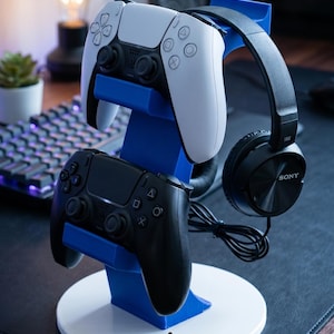 May include: A blue and white 3D-printed stand for gaming controllers and a headset. The stand holds a white and a black controller, and a black headset. Text at the top says "STOP THE DESK MESS!" and at the bottom "3D PRINTED!"