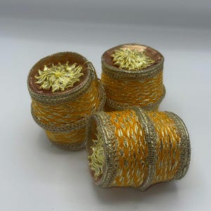 May include: Three cylindrical napkin rings featuring a bright orange woven texture with metallic gold thread accents. Each ring is topped with a decorative gold floral embellishment and finished with a gold metallic trim along the edges.