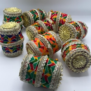 Può includere: Una collezione di oggetti decorativi cilindrici colorati. Ogni pezzo presenta ricami intricati in rosso, verde, giallo e arancione, accentuati da finiture dorate. Le parti superiori e inferiori sono decorate con abbellimenti dorati.