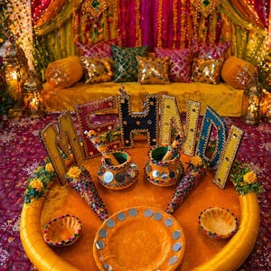 May include: A colourful, festive scene with the word "Mehndi" in illuminated letters. The setting includes a decorated tray with decorative items, pots, and a plate, all in shades of orange and gold. The background features colourful fabrics and pillows.
