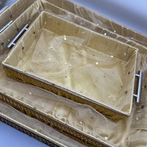 May include: A set of three rectangular decorative trays in varying sizes. The trays are a light gold colour with silver handles and are lined with a sheer, gold-toned fabric dotted with gold sequins. The trays are nested together.