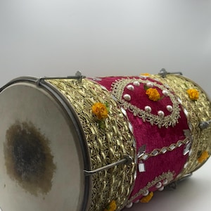 May include: A dhol drum featuring a deep magenta velvet body adorned with intricate gold metallic lace trim, small white faux pearls, reflective mirror accents, and small yellow fabric flowers, secured with metal tension rods and a drumhead.