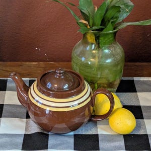 May include: A brown ceramic teapot with yellow stripes, a lid, and a handle sits on a black and white checkered cloth. A green glass vase with green leaves and two yellow lemons are also on the cloth.