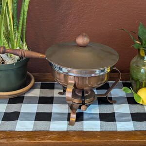 May include: A copper and stainless steel fondue pot with a wooden handle and lid. The pot sits on a stand with a burner. A potted plant and a green vase with greenery are on either side of the pot. Two lemons are in front of the vase.